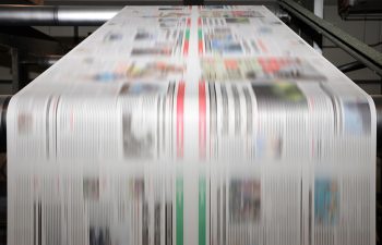 A large offset printing press running a long roll off paper over its rollers at high speed.